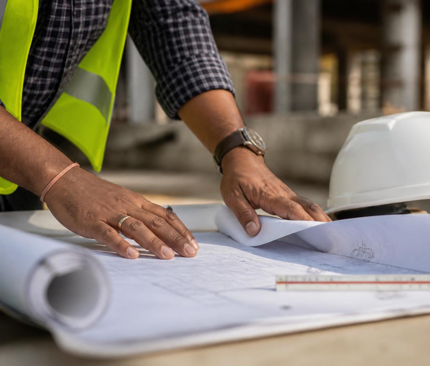 Site engineer in a safety vest reviewing rolled architectural drawings on a table beside a hard hat
