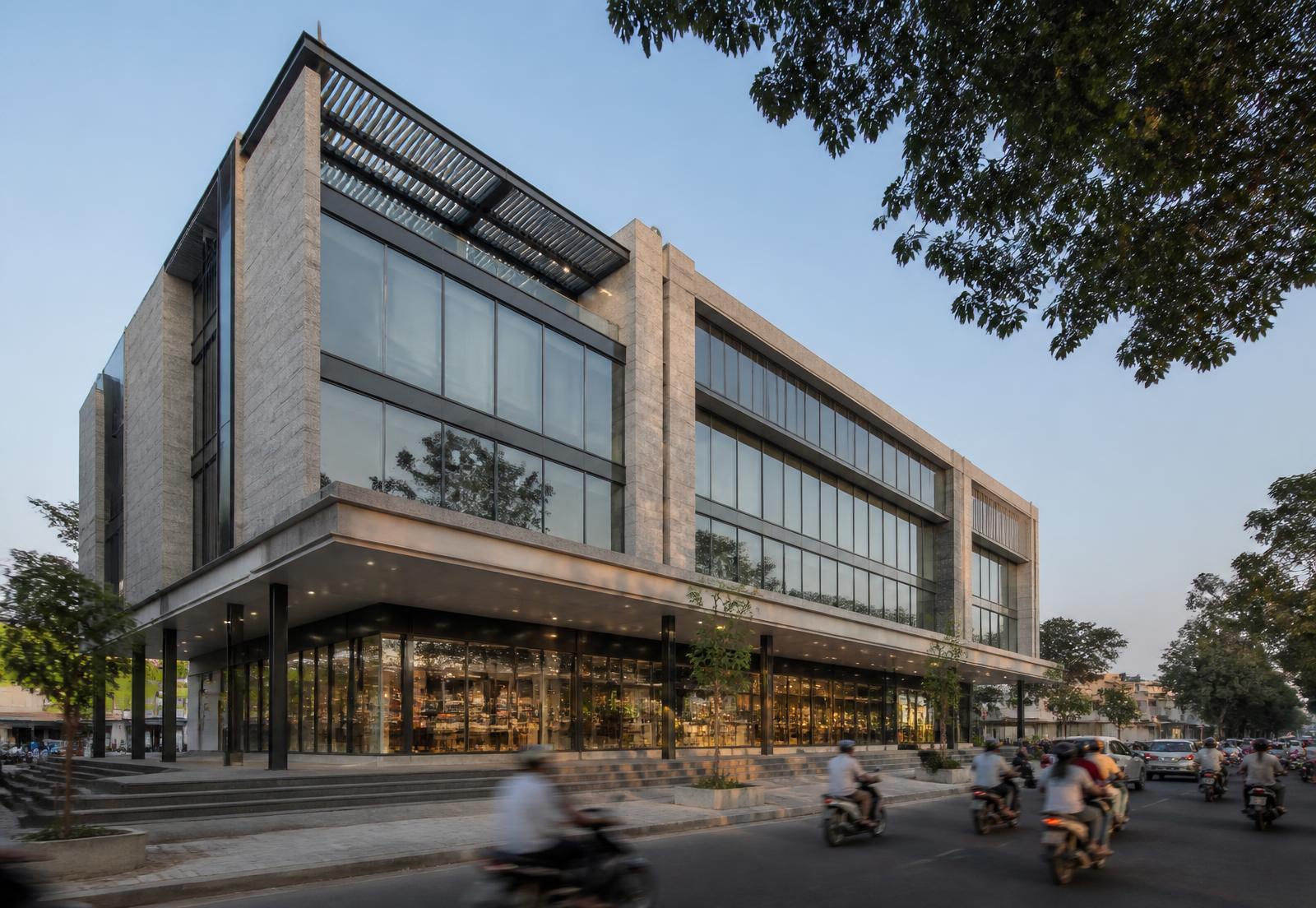 Modern mixed-use commercial building in stone and glass with shopfronts under a deep canopy beside a busy street